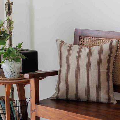A square cushion cover with vertical brown and beige stripes, resting on a dark wood bench in a cozy, rustic room setting.