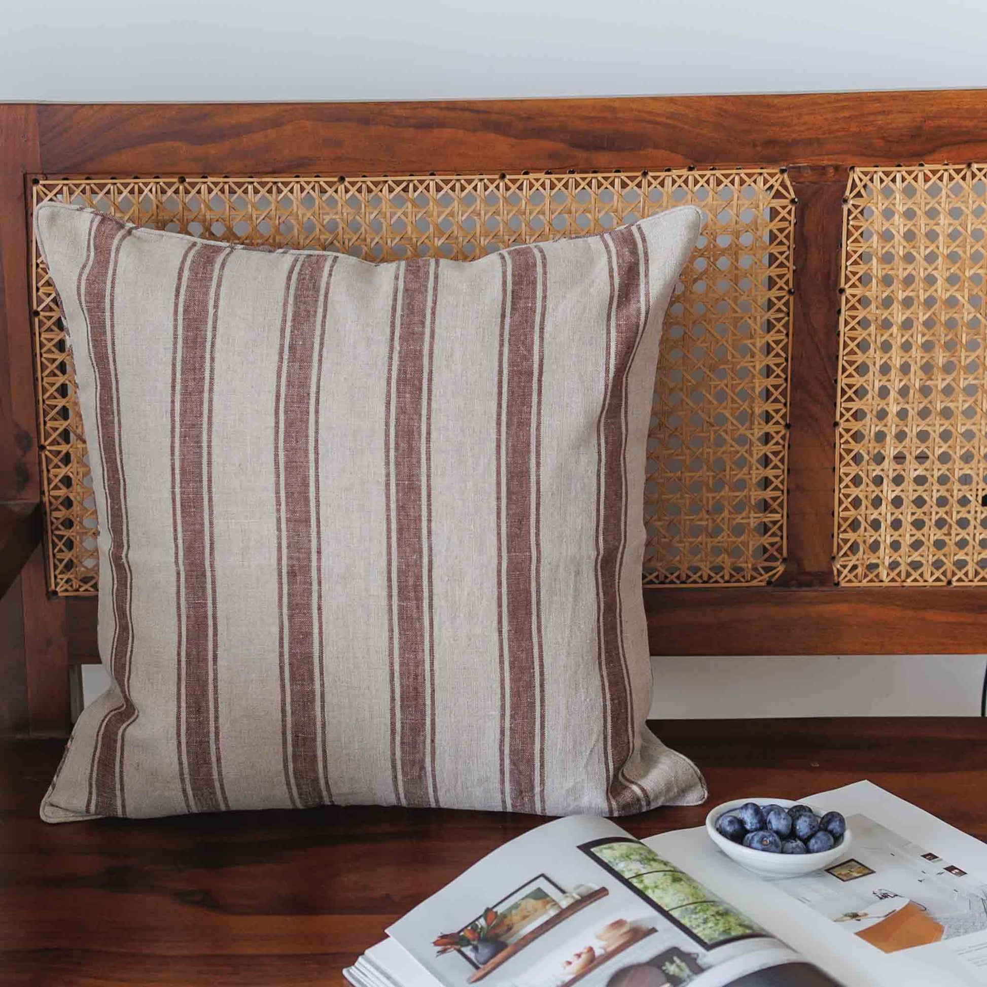 A square throw pillow with vertical brown and beige stripes, styled on a dark wooden bench with woven cane panels, next to an open magazine and a small bowl of blueberries.