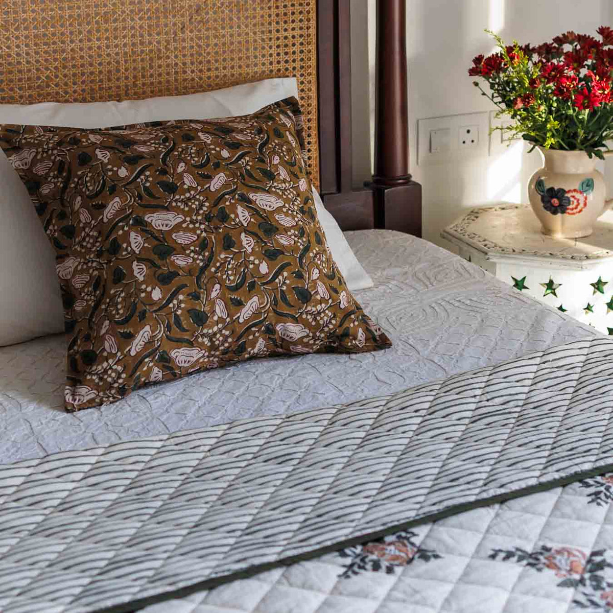 A block print decorative cushion cover with a brown floral pattern, styled on a bed with a striped floral quilt.