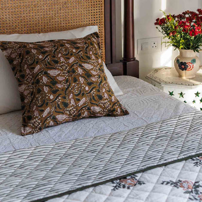 A block print decorative cushion cover with a brown floral pattern, styled on a bed with a striped floral quilt.