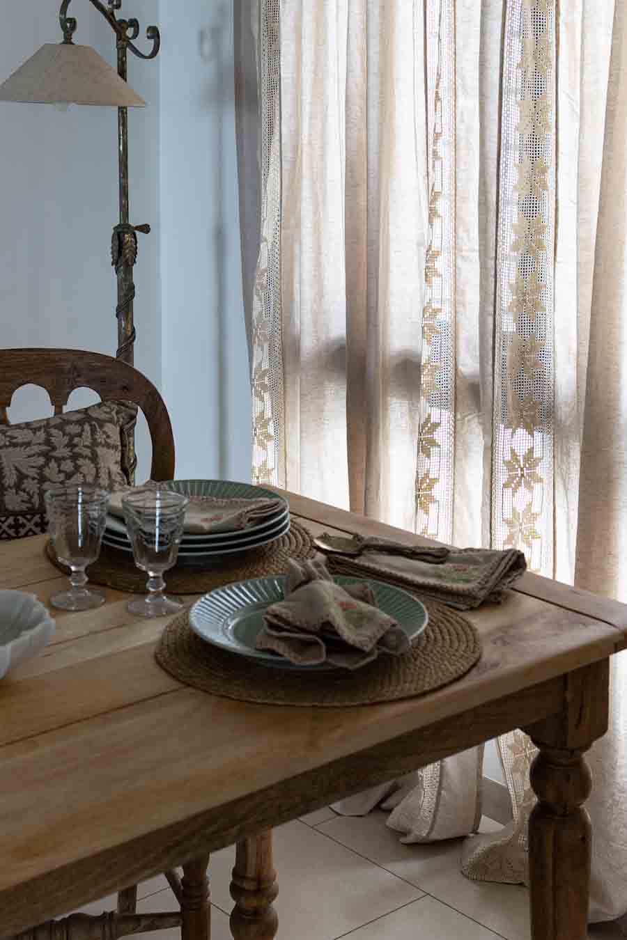 A rustic dining setting with plates, woven placemats, folded linen napkins, in a softly lit room with sheer lace curtains.
