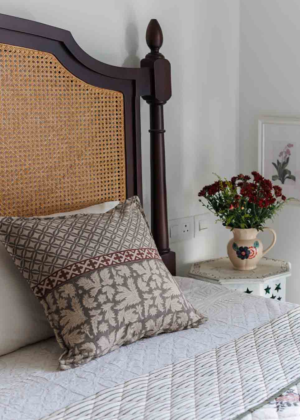 A bedroom dressed in block print quilt and a block print cushion cover with white bedside table with a pitcher of red flowers.