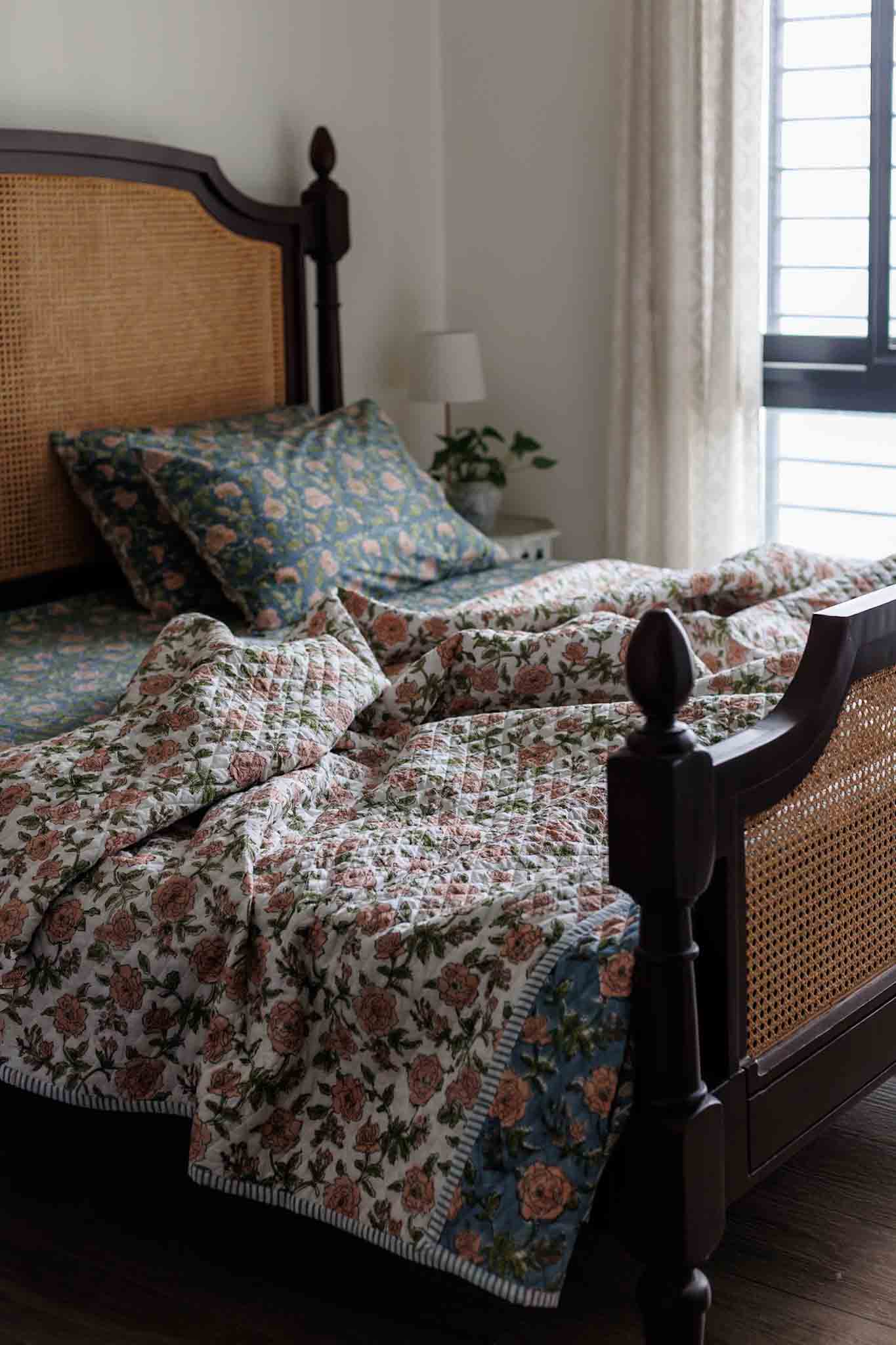 A floral quilted bedding set with matching pillowcases on a dark wooden bed with a woven cane headboard, styled in a cozy bedroom setting.