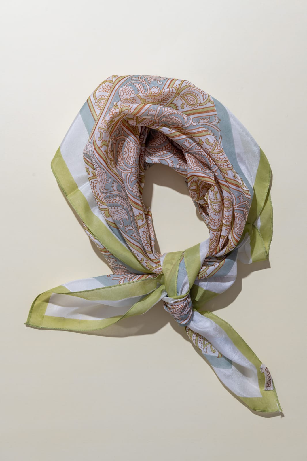 A square silk scarf with a complex paisley and stripe pattern in muted tones of sage green, dusty blue, beige, and white, tied in a knot and displayed on a warm off-white background.