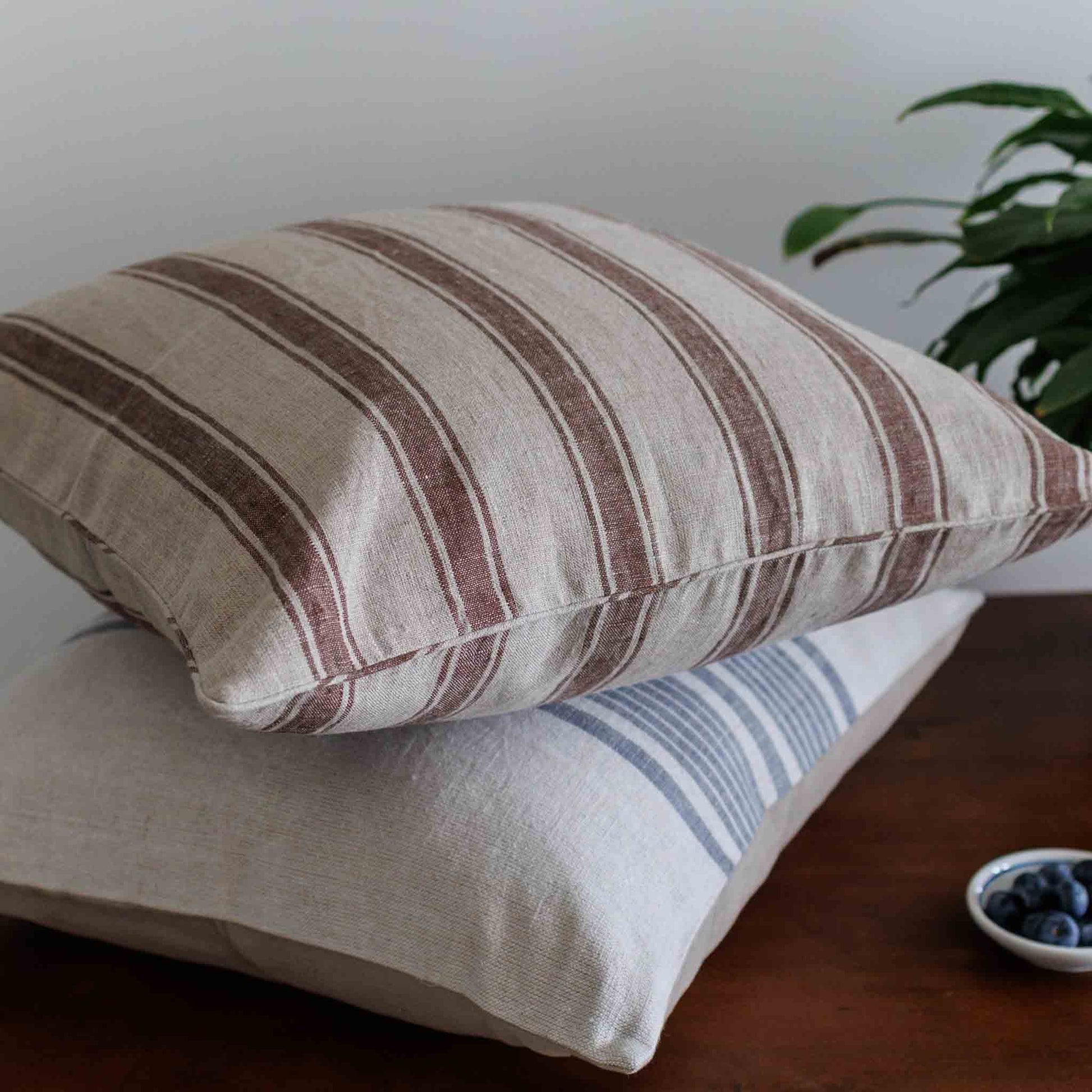 Two striped linen cushion covers stacked on a wooden table, with a small bowl of blueberries and a green plant in the background.