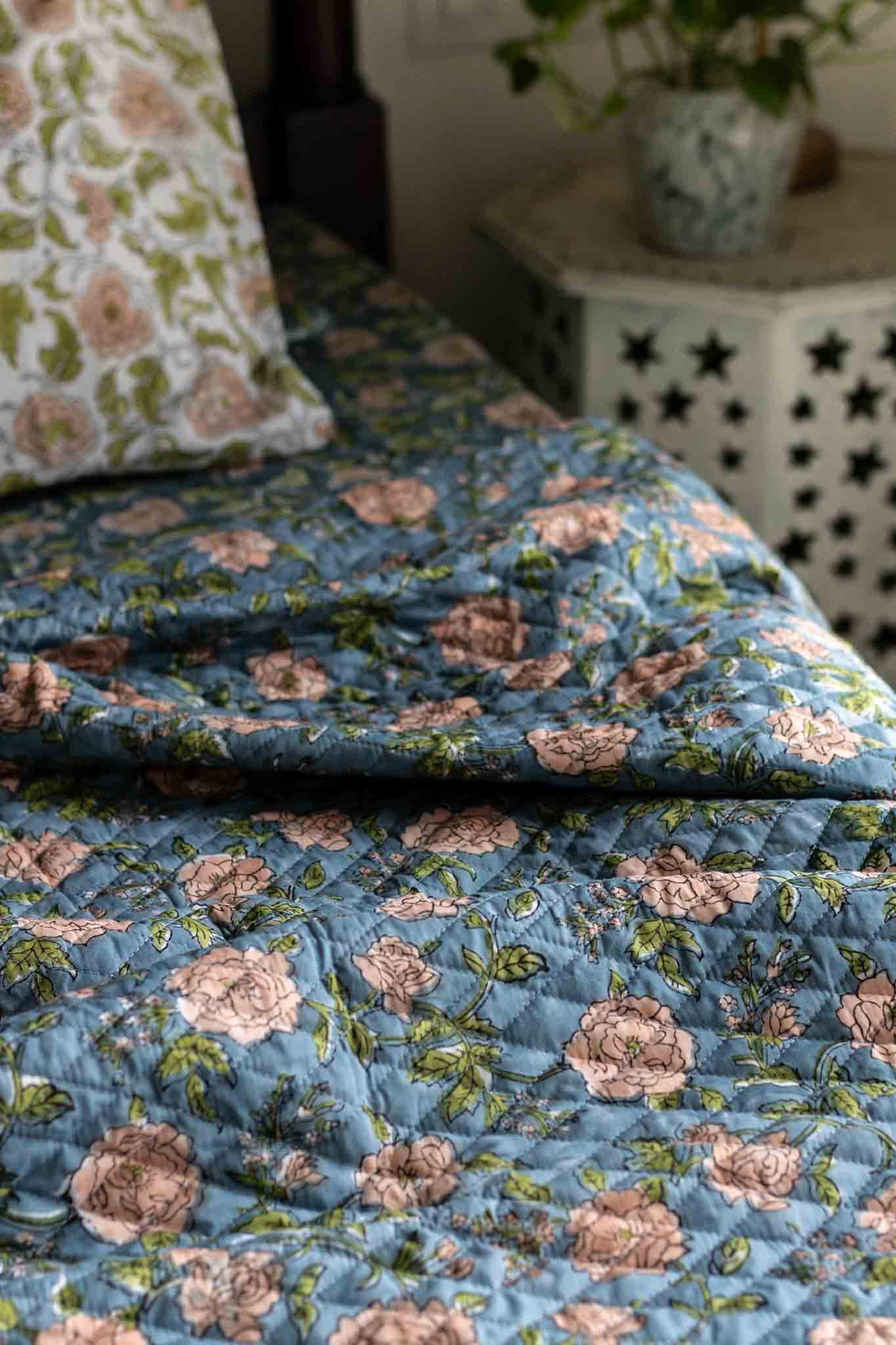 A close-up of a quilted blue floral comforter or bedspread with pink roses and green leaves, styled on a bed with a matching pillow in a softly lit bedroom.