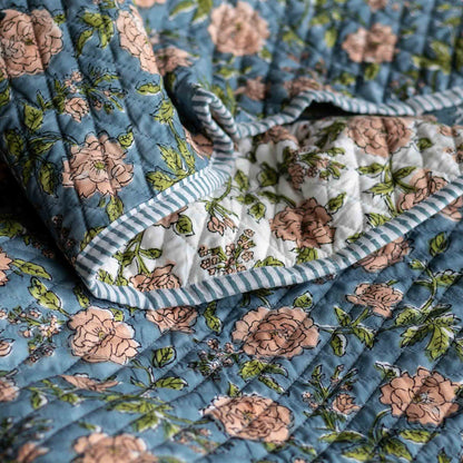 A close-up of a quilted blanket with a dusty blue floral print, featuring pink roses, green leaves, and striped blue and white borders.