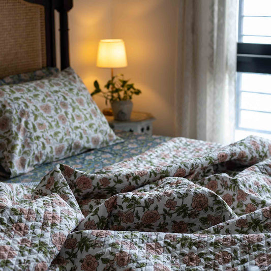 A quilted floral bedding set with matching pillowcases on a bed in a cozy bedroom setting, featuring a warm glowing bedside lamp and window curtains.