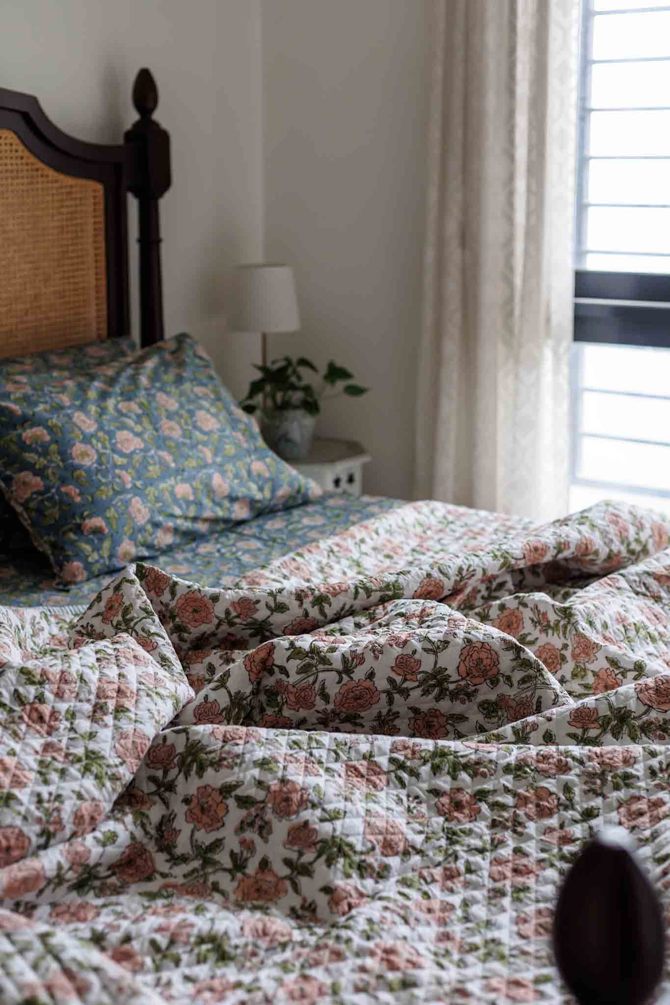 A close-up of a quilted bedding set with a floral pattern in dusty pink, sage green, and blue on a white background, styled on a bed in a softly lit bedroom.