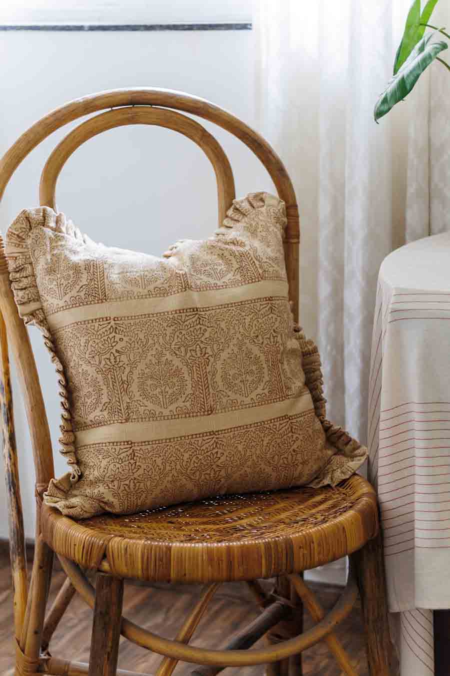 A square rust and beige block print throw pillow with ruffled edges, resting on a woven rattan chair in a softly lit room.