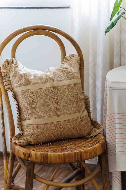 A square rust and beige block print throw pillow with ruffled edges, resting on a woven rattan chair in a softly lit room.