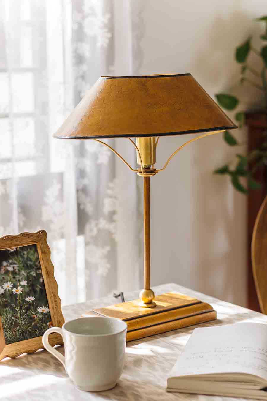 A Study table lamp with a textured bronze shade and brass-toned metal stand, styled on a  tabletop next to a white ceramic mug, an open book, and a small framed picture in a softly lit room.