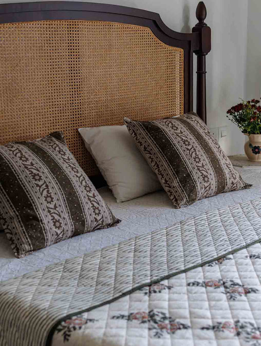 A  bedroom styled in a woven rattan bed with a green and brown pattern block print cushion covers and a floral quilted bedspread.