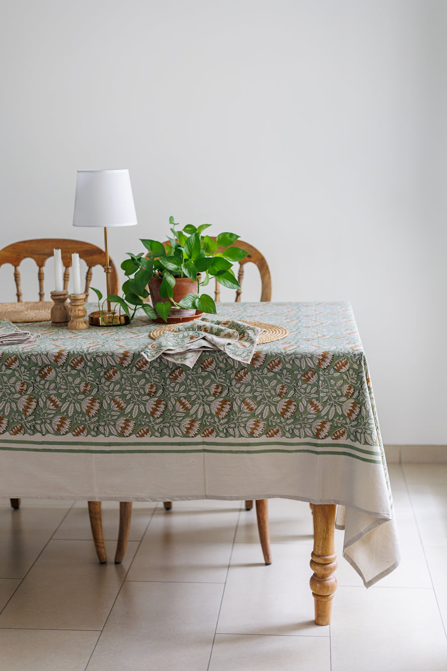 Table with a patterned Sage green tablecloth, plant, and lamp in a room.